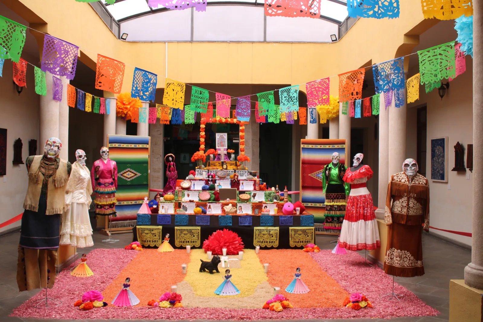 Altar de muertos al aire libre con papel picado, tapete de aserrín de colores y catrinas con trajes típicos mexicanos