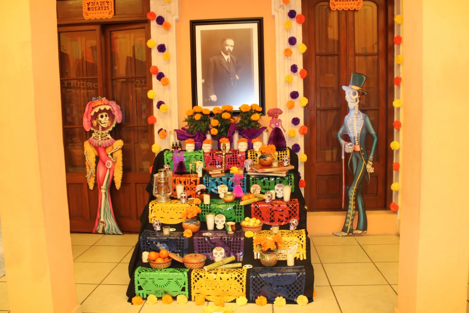 Altar de muertos escalonado con papel picado de colores, cempasúchil, calaveritas y un retrato de Francisco I. Madero, flanqueado por figuras de catrina y catrín
